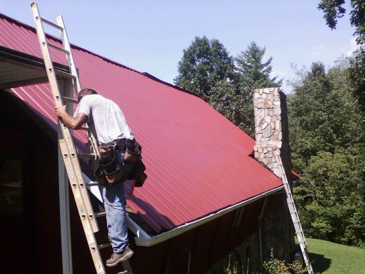 Roofer on a ladder beside a red metal roof and stone chimney — Soffit & Fascia Repair by Roofing Brothers in Mount Summit